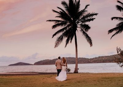 Bride and Groom standing under a palm tree at a pink sunset overlooking the ocean, at Paradise Cove Resort in the Whitsundays